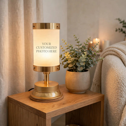 A brass table lamp with a glowing frosted cylindrical shade stands on a wooden side table next to a potted eucalyptus plant in a cozy room setting. The lamp has gold-toned metal accents and a circular base.