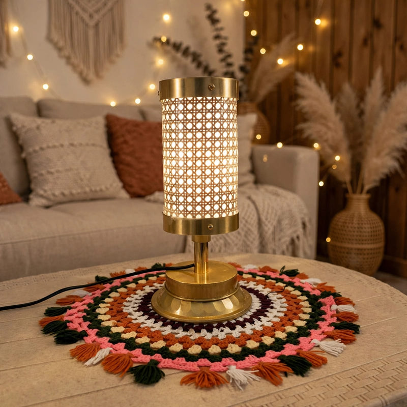 A brass table lamp with a cylindrical screen featuring a geometric cutout pattern, glowing with warm light. It sits on a round, colorful crocheted doily with tassels on a light-colored wooden table in a cozy, warmly lit living room setting.