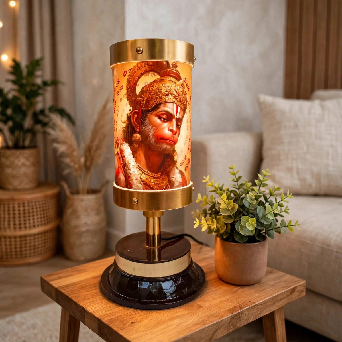 A decorative table lamp featuring a cylindrical amber-toned shade printed with an illustration of Hanuman, set on a wooden side table in a cozy living room with a sofa and potted plants in the background.