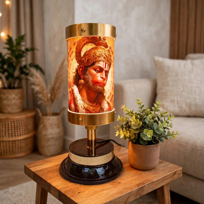 A decorative table lamp featuring a cylindrical amber-toned shade printed with an illustration of Hanuman, set on a wooden side table in a cozy living room with a sofa and potted plants in the background.