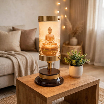 A decorative table lamp with a glowing Buddha figurine inside a clear cylindrical shade, set on a wooden side table in a softly lit living room. The lamp has a brass-toned metal frame and base with a dark round pedestal.